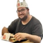 Subject | Rhodes Sea Lines Man wearing a paper Burger King crown, glasses, and a dark t-shirt, sitting at a table with a fast-food tray and bag.